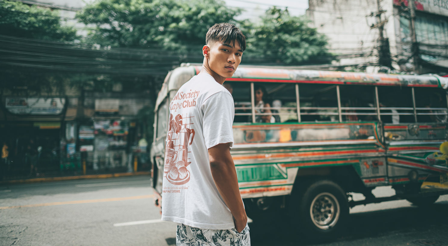 A filipino boy walking in the streets of manila with a jeepney behind him wearing a 4AM Soceity T Shirt in white